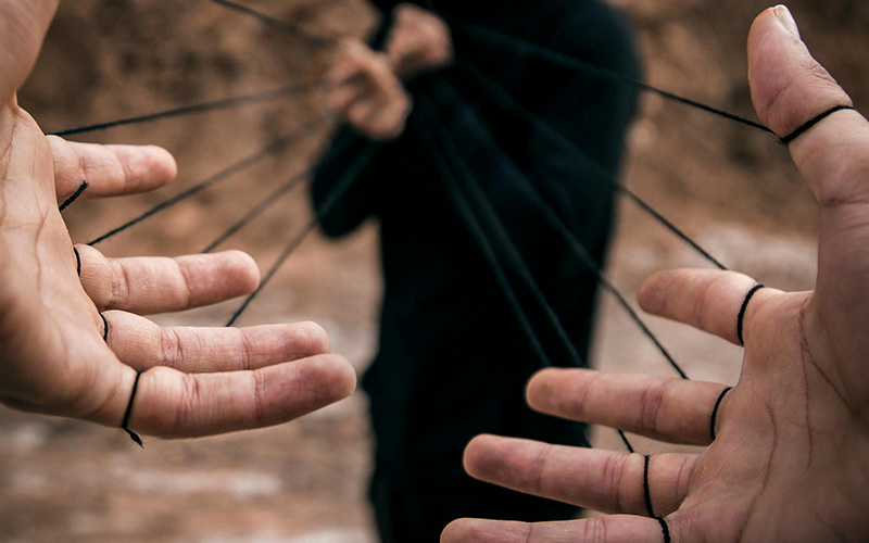 Mãos de uma pessoa com fios atados em cada dedo puxados por uma outra pessoa desfocada à distância para simbolizar a manipulação pelo algoritmo nas redes sociais