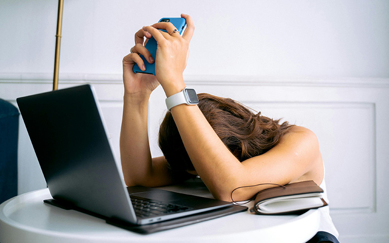 Mulher com ar desesperado deitada sobre o computador portátil e com o telemóvel na mão, a simbolizar a fadiga digital